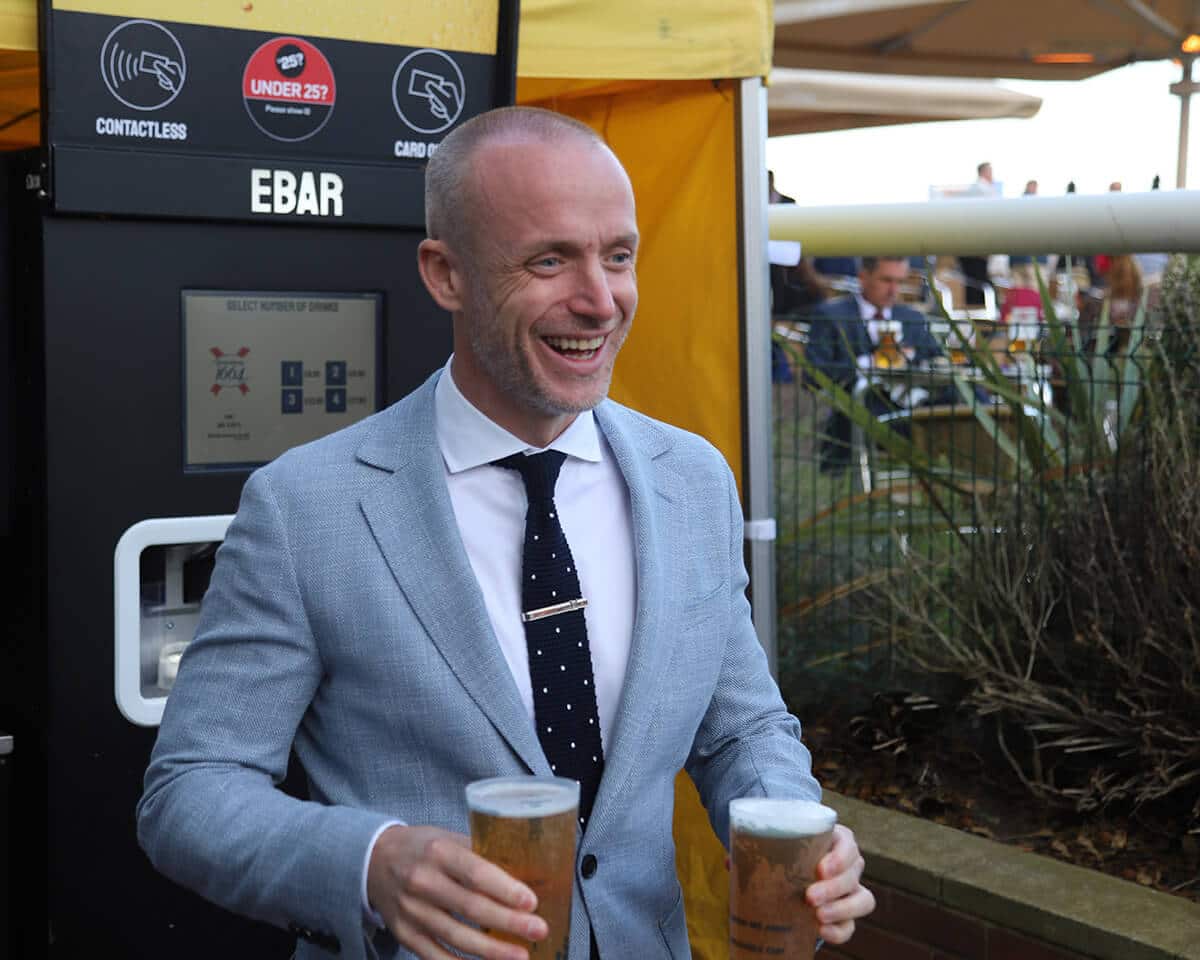 A smiling man in a suit walks away from an EBar unit, holding two pints of lager