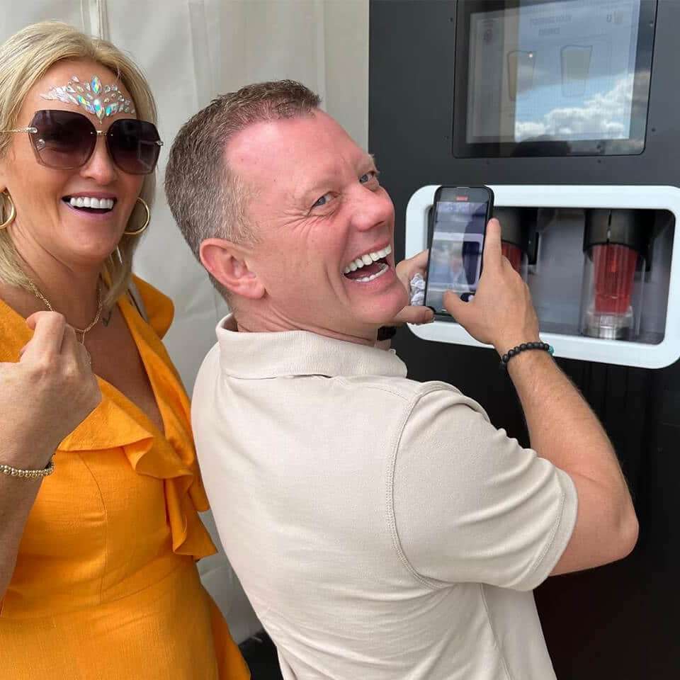 A woman and a man laugh to the camera as they use a phone to film a mobile EBar pouring their pints of pink cider