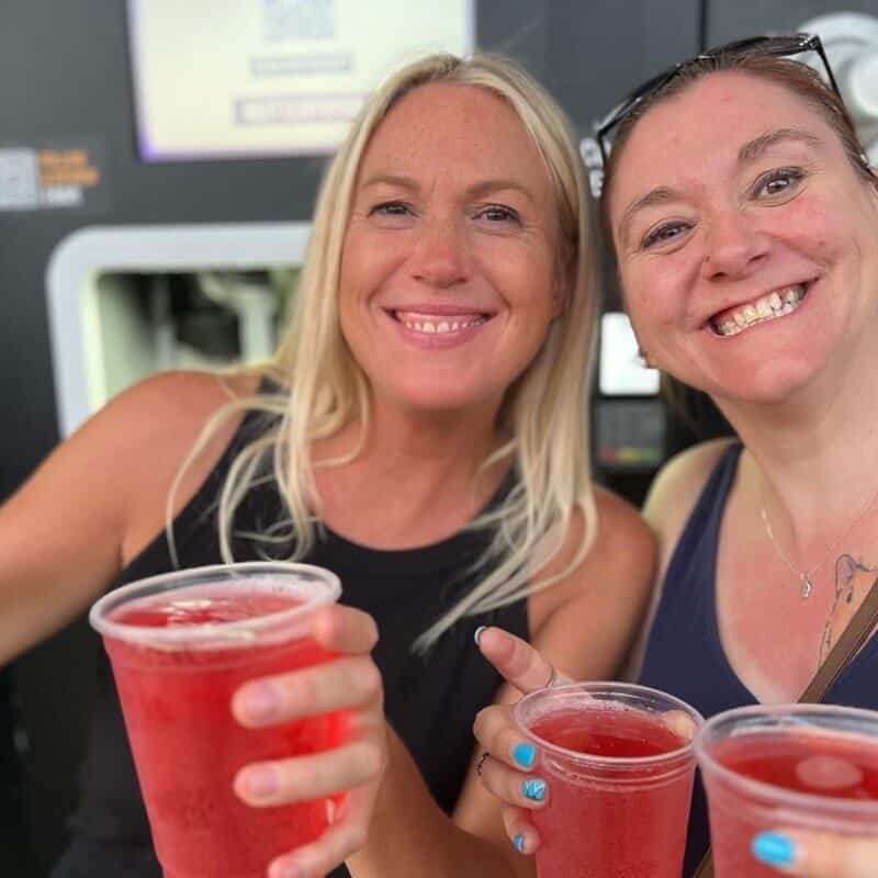 Two women smile for the camera while showing off the 4 pints of pink cider they have just received from the EBar units in the background