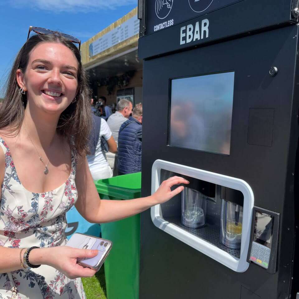 A woman with sunglasses on her head pours herself two pints of lager from a mobile EBar automated beer kiosk