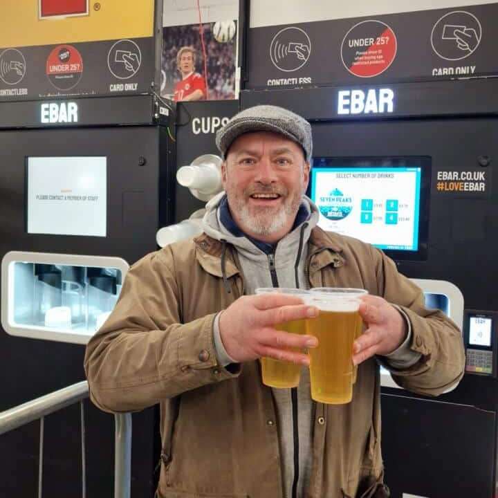 A man in a flat cap holds three pints that he just poured from the mobile EBar unit in the background