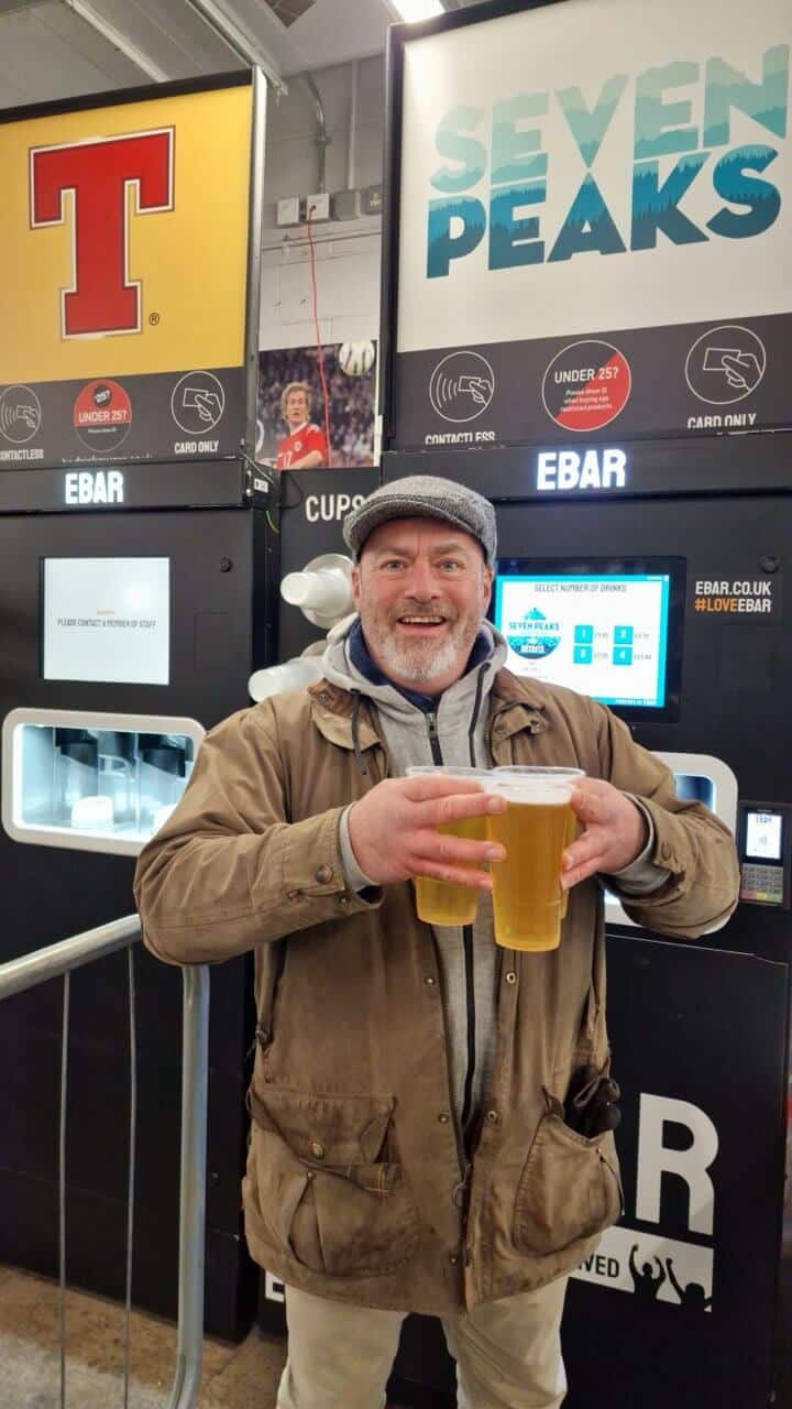 A man in a flat cap holds three pints that he just poured from the mobile EBar unit in the background