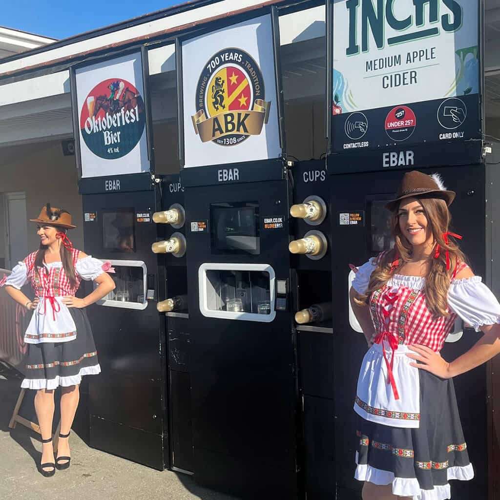 Two women in traditional Oktoberfest outfits stand smiling in front of 3 mobile EBar units
