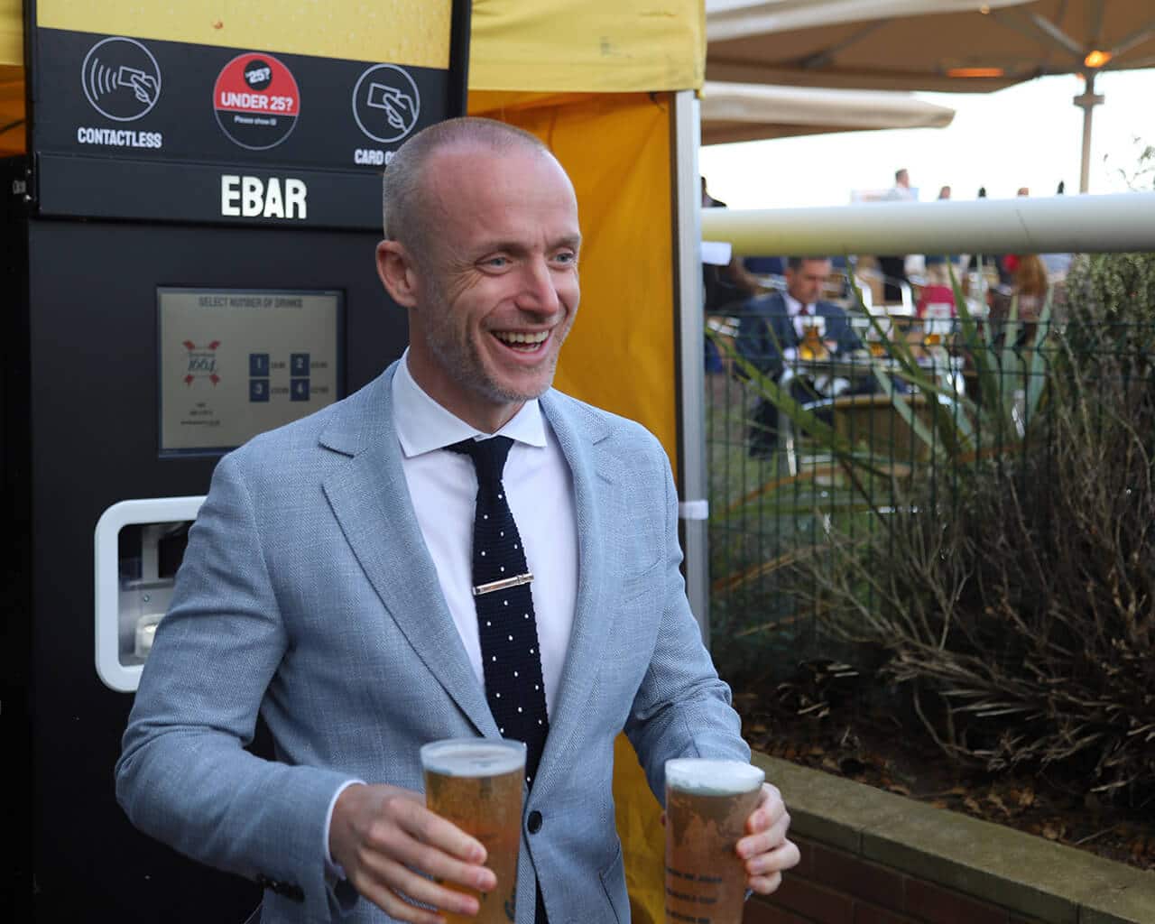 A smiling man in a suit walks away from an EBar automated beer kiosk , holding two pints of lager
