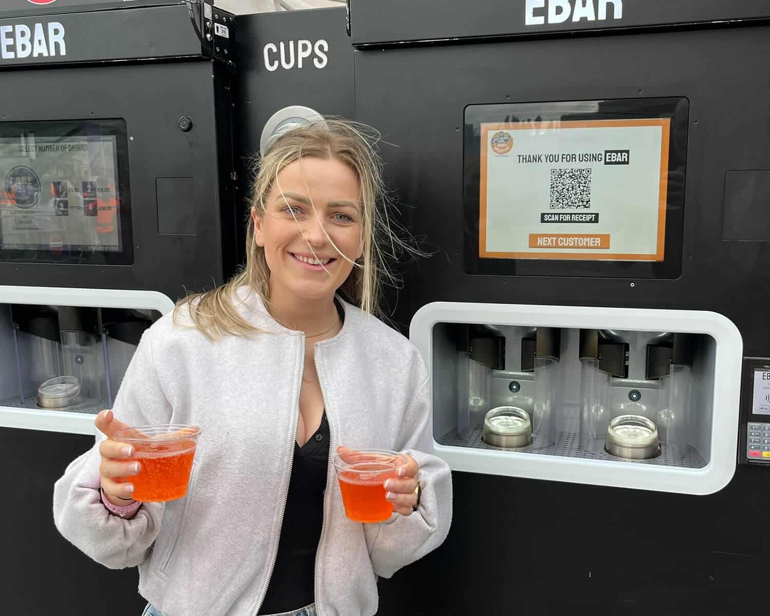 A smiling woman holds up two cocktails she just purchased from the EBar unit behind her