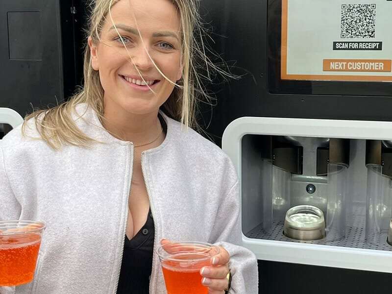 A smiling woman holds up two cocktails she just purchased from the EBar unit behind her