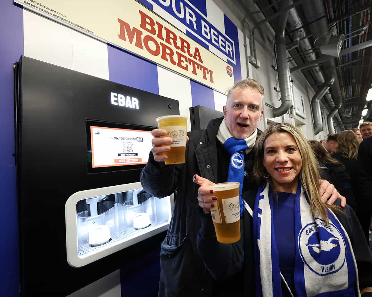 Two football fans smile to the camera after using the Beerwall behind them at the Amex Stadium, Brighton and Hove Albion FC