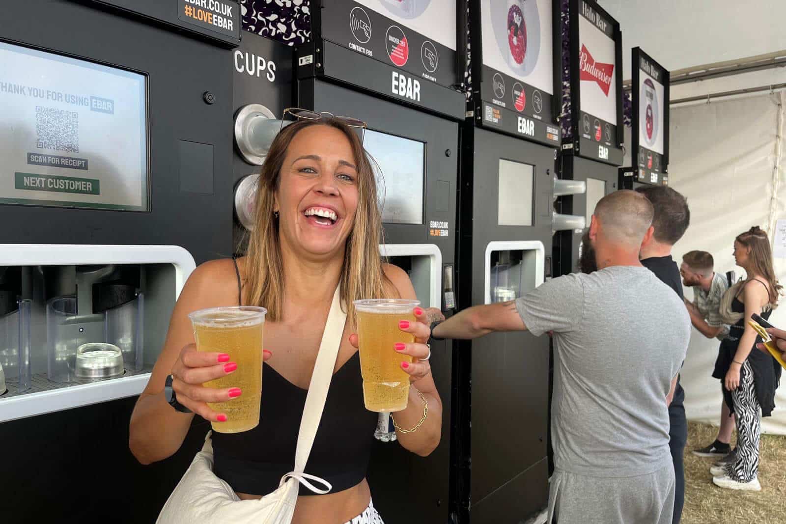 A laughing woman holding 2 pints of Budweiser lager with multiple mobile EBar units behind her